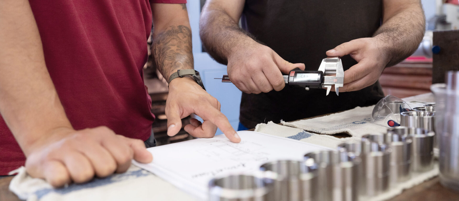 Zwei Mitarbeitende, die sich austauschen. Eine Person zeigt auf einen Plan, die andere miss ein Metallteil ab.